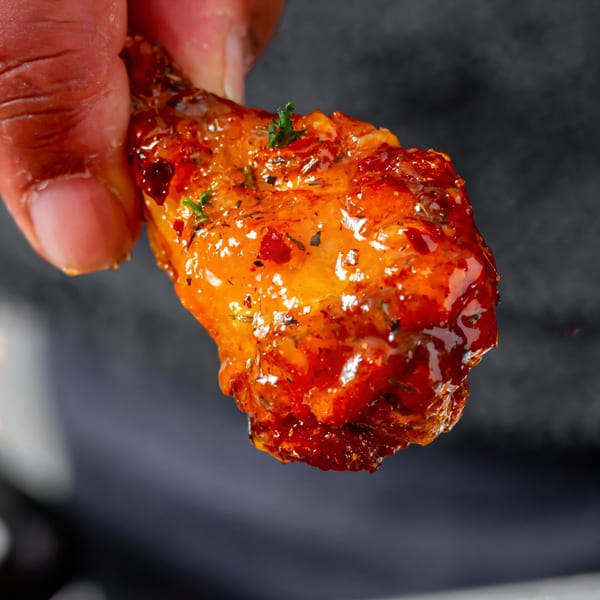 A hand holds up a glistening, saucy chicken wing, garnished with herbs, from Next Phaze Cafe.