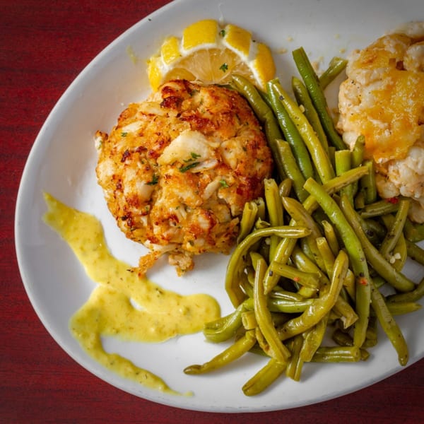 A crab cake with green beans, cheesy cauliflower, lemon slices, and a yellow sauce on a white plate.