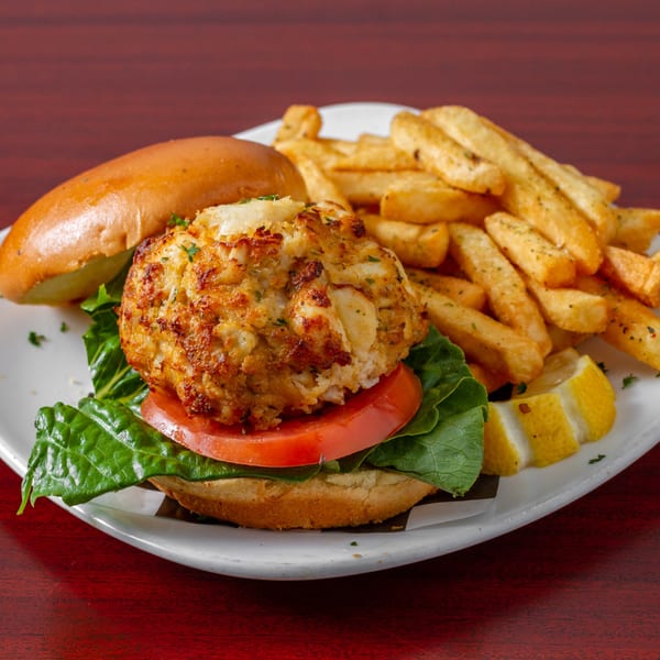 A crab cake sandwich on a bun with lettuce and tomato, served with french fries and lemon wedges.